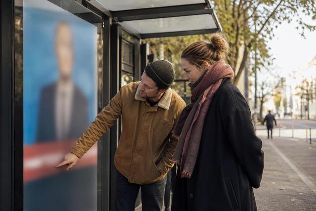 Eine Bürgerin zeigt ihrem Begleiter den Hinweisbereich eines politischen Werbeplakats an einer Haltestelle.