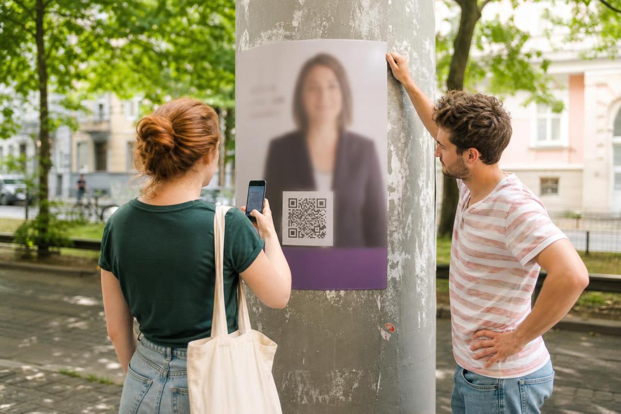 Zwei Ehrenamtliche prüfen den QR-Code eines Wahlplakats mit einem Smartphone.