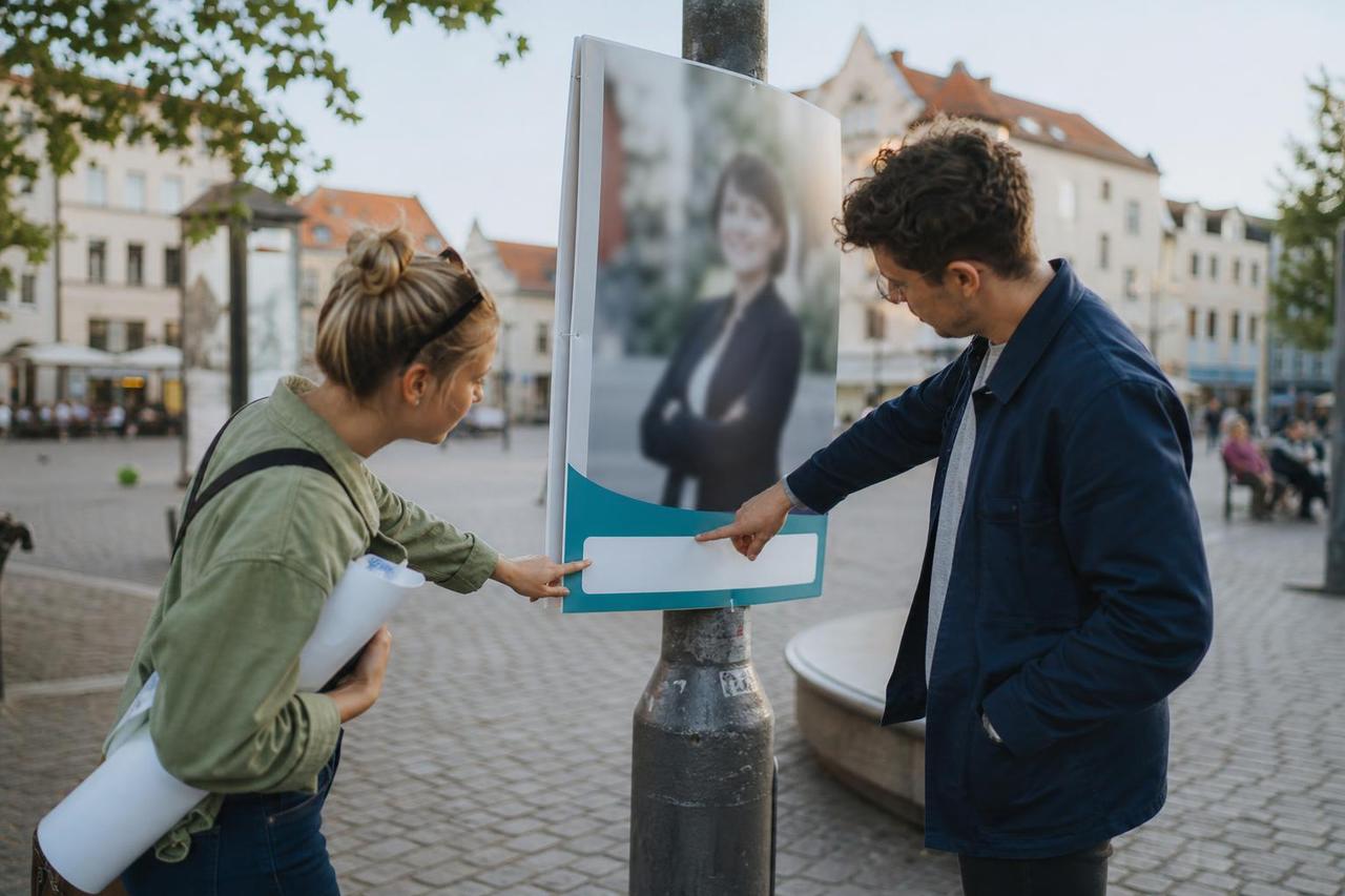 Zwei Ehrenamtliche prüfen das Informationsfeld eines politischen Plakats auf einem Marktplatz.