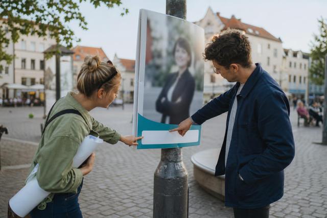 Zwei Ehrenamtliche prüfen das Informationsfeld eines politischen Plakats auf einem Marktplatz.