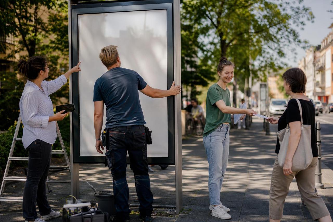 Ein Wahlkampfteam prüft eine Werbefläche, während eine Helferin nebenan einen Flyer verteilt.