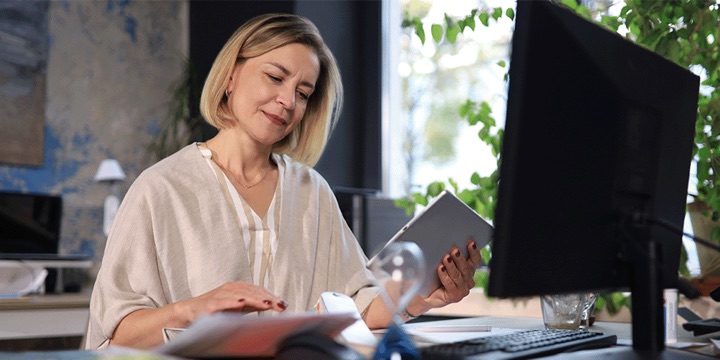 Frau am Arbeitsplatz mit Tablet in der Hand und Bildschirm auf dem Schreibtisch